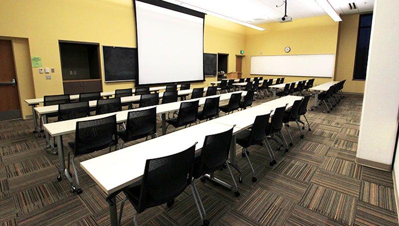 Photo of a reservable space containing screen and projector, white board, table rows with chairs in classroom setting.