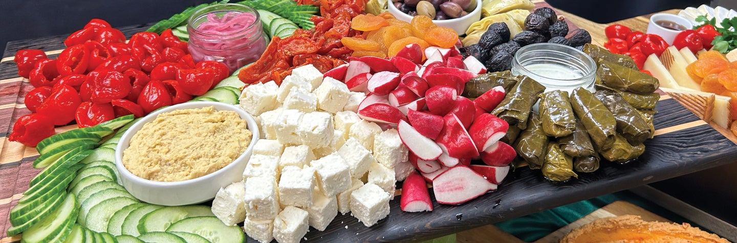 Appetizer tray featuring cheeses, cucumbers, radishes, grape leaves, dates, sun-dried tomatoes, peppers