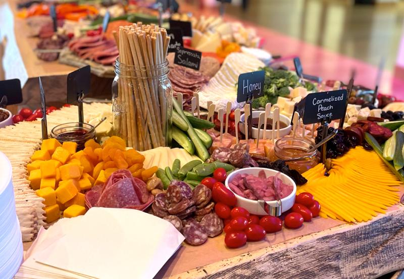 Photo of charcuterie table containing variety of cheeses, salami, tomatoes, and vegetables.